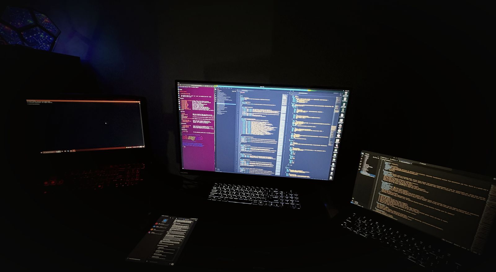 My desk at night: a center monitor and two laptops full of code, a phone, and a glowing dodecahedron lamp